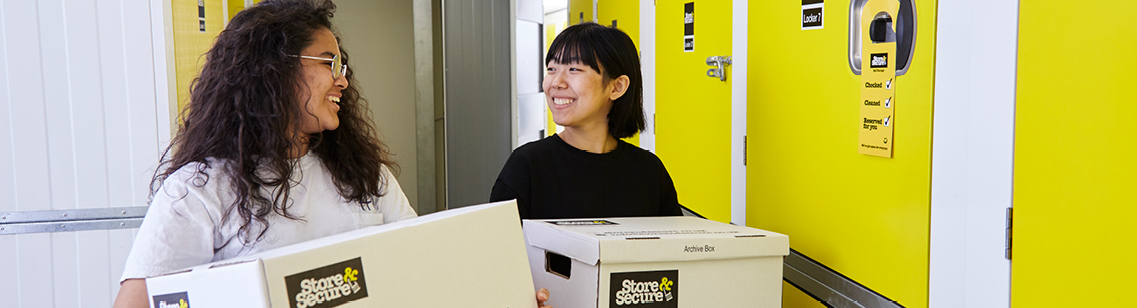 Young happy pair carrying Store & Secure boxes through a holiday storage unit