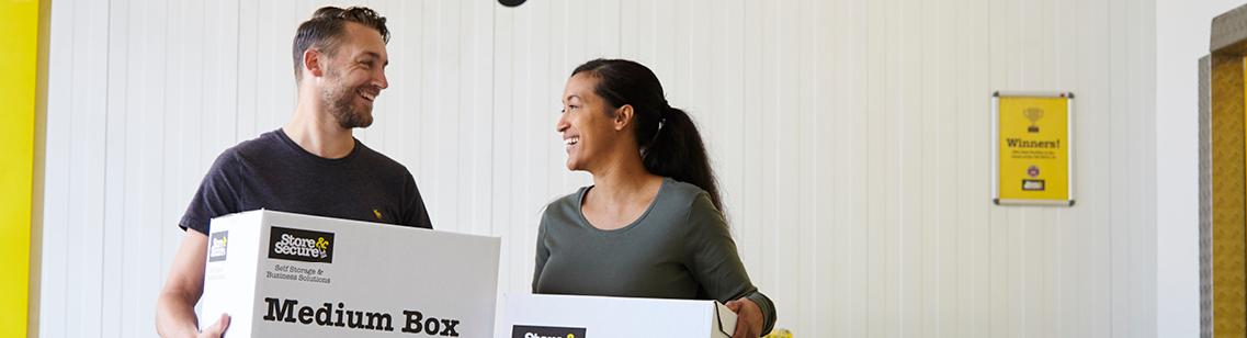 Young man and woman smiling at each other while holding Store & Secure storage boxes in a self-storage unit